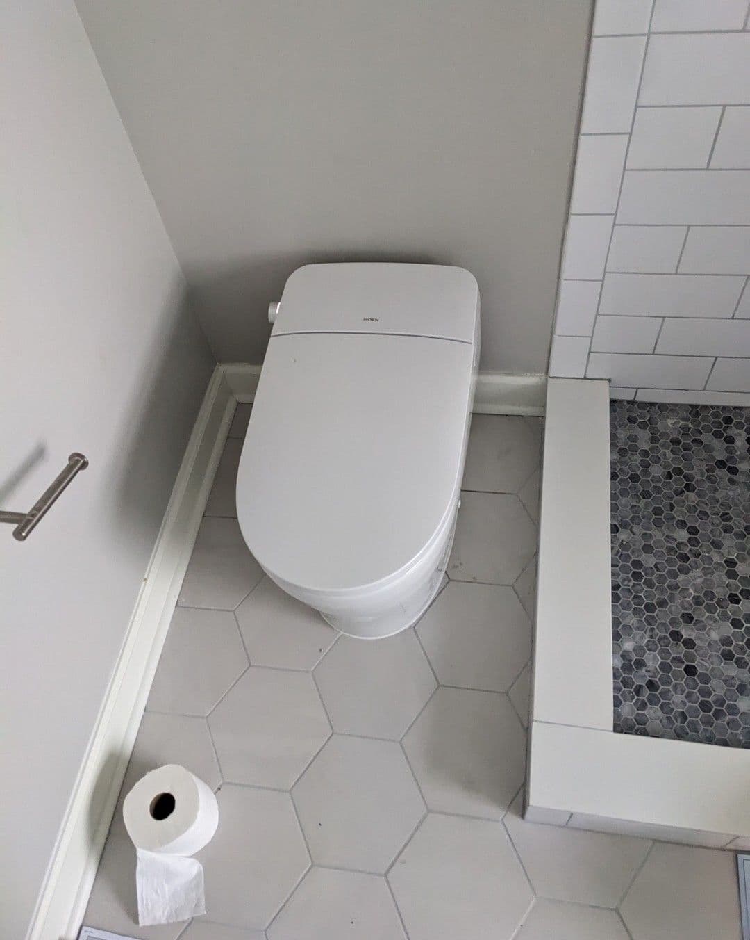 Modern white toilet in a minimalistic bathroom with gray hexagonal tiles and a shower area.