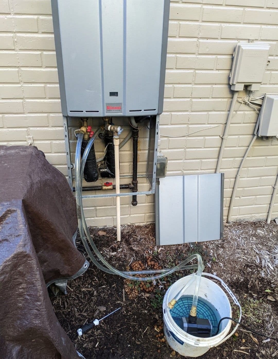 Tankless water heater installation with connected hoses and water bucket on the ground.