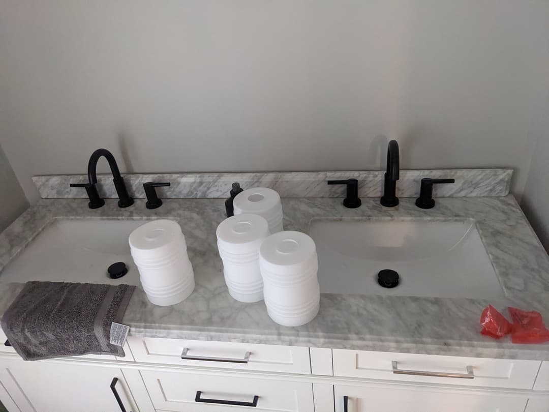 Dual bathroom sinks with black faucets, marble countertop, and stacked white cups.