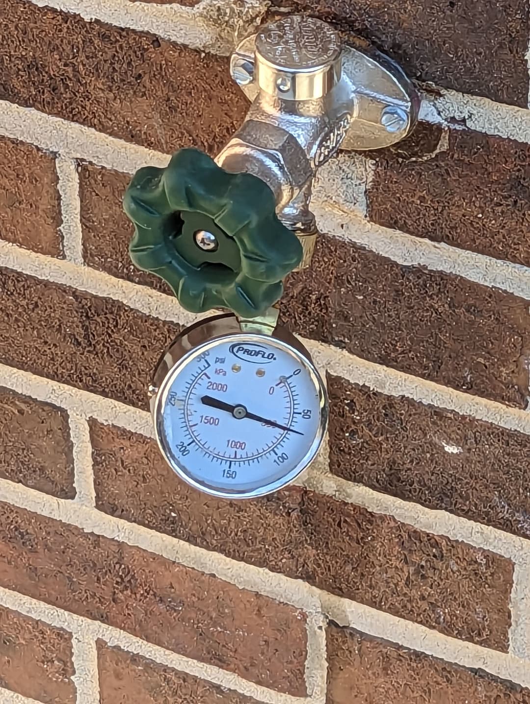 Water pressure gauge with green valve mounted on a brick wall.