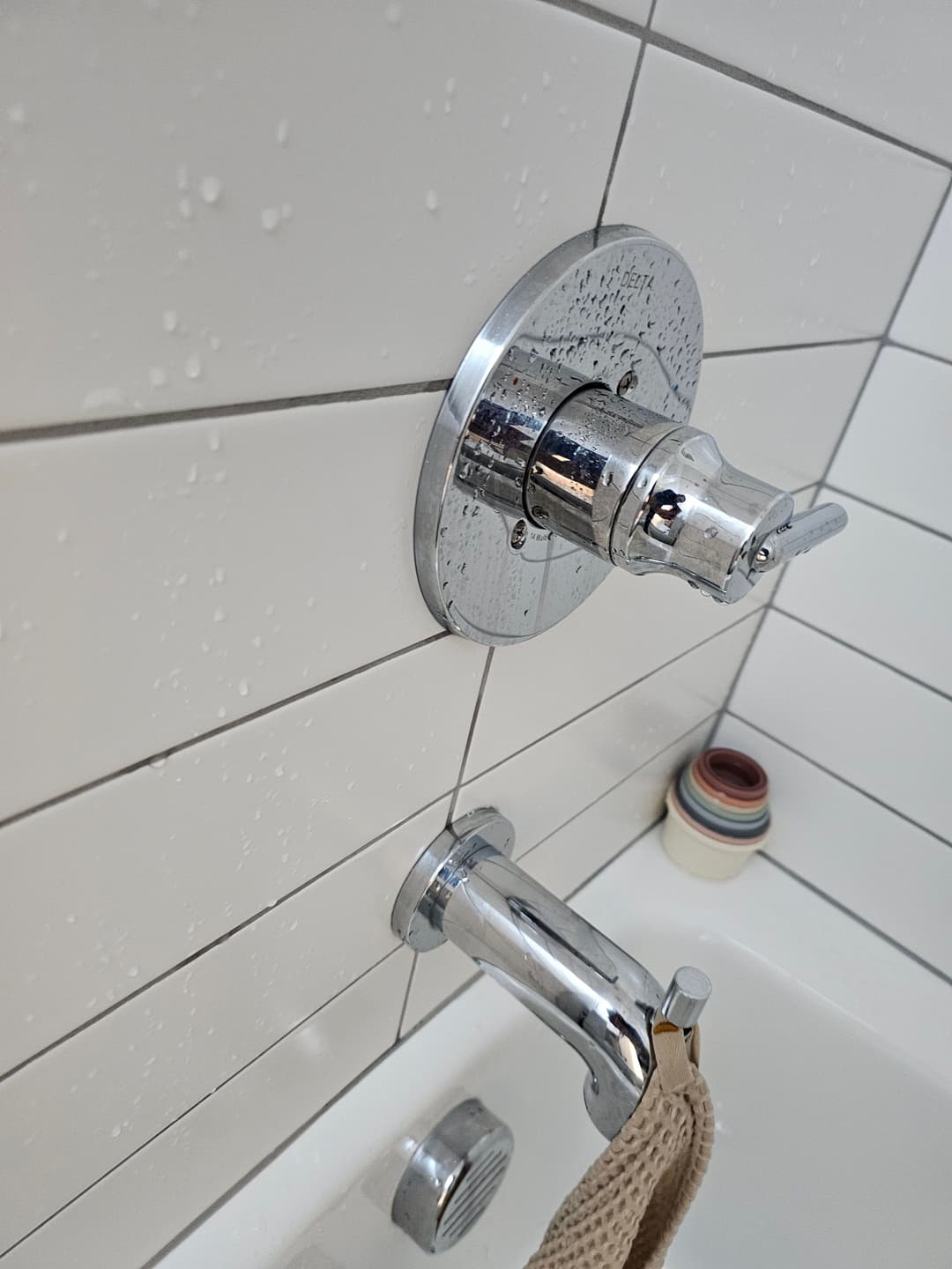 Chrome shower faucet with wall mount, water droplets, and colorful shampoo bottles in the background.