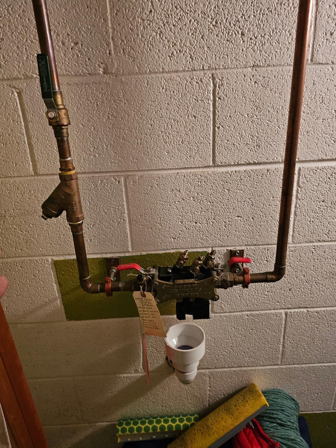 Piping and valve system mounted on a wall, with a white cup and labels attached.