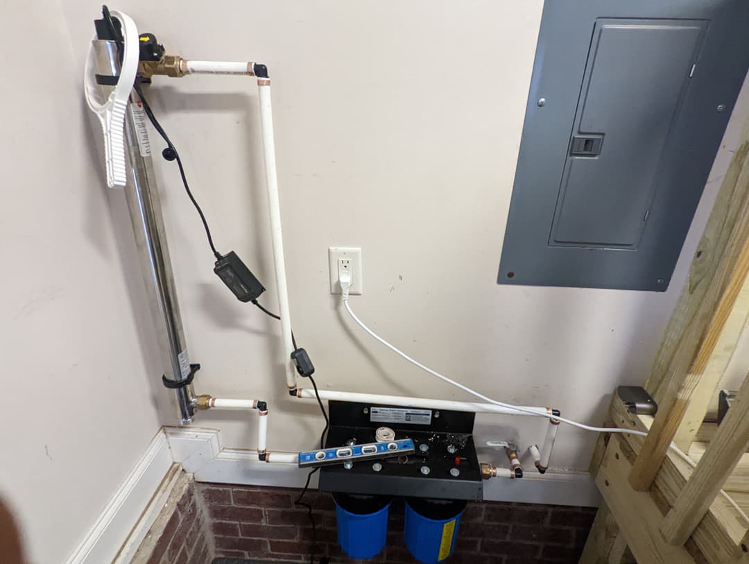 Home water filtration system with white PVC pipes, connected to a control panel and power outlet.