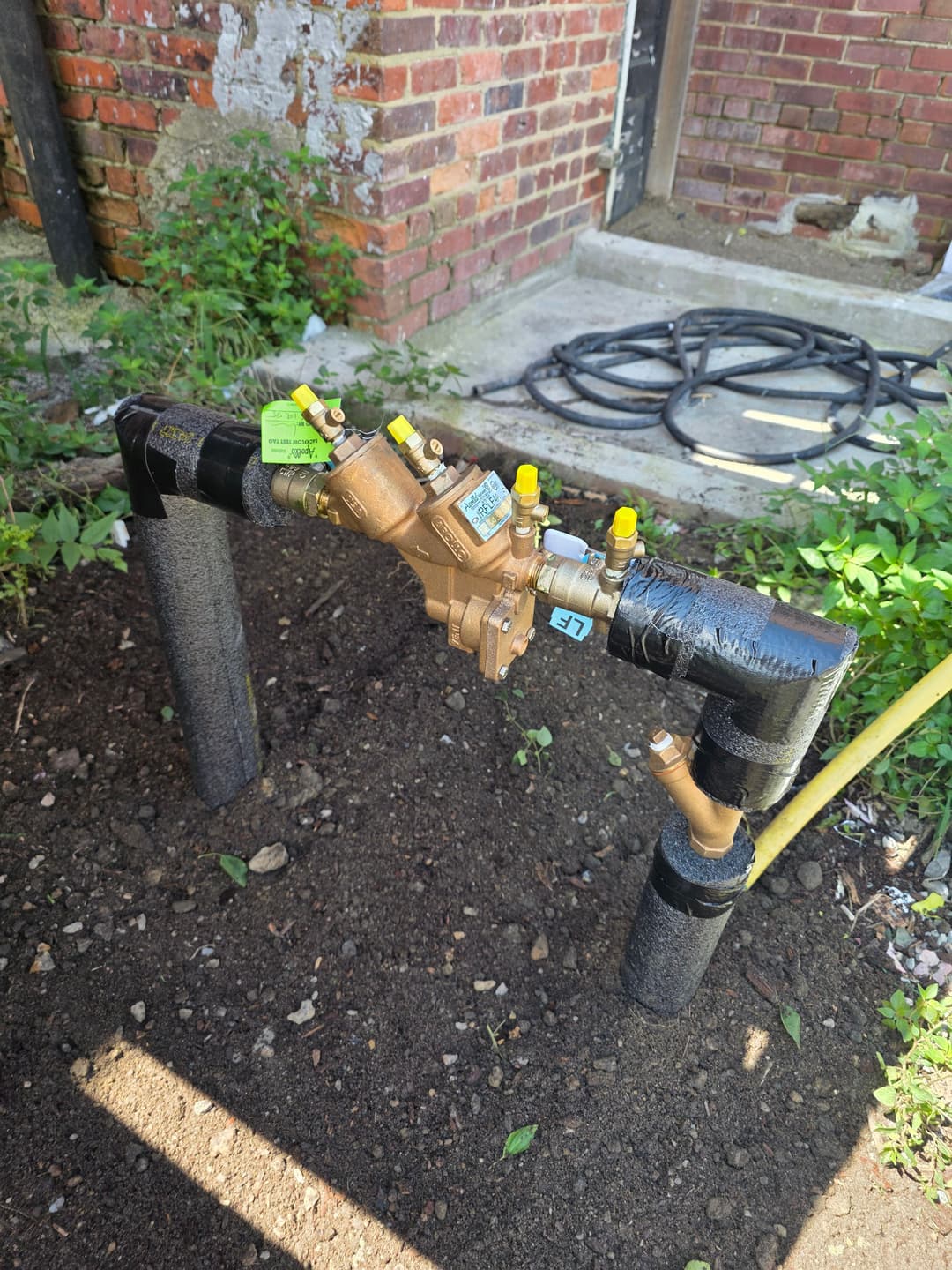 Water pressure regulator and pipes installed in a garden setting, with insulation on pipes.