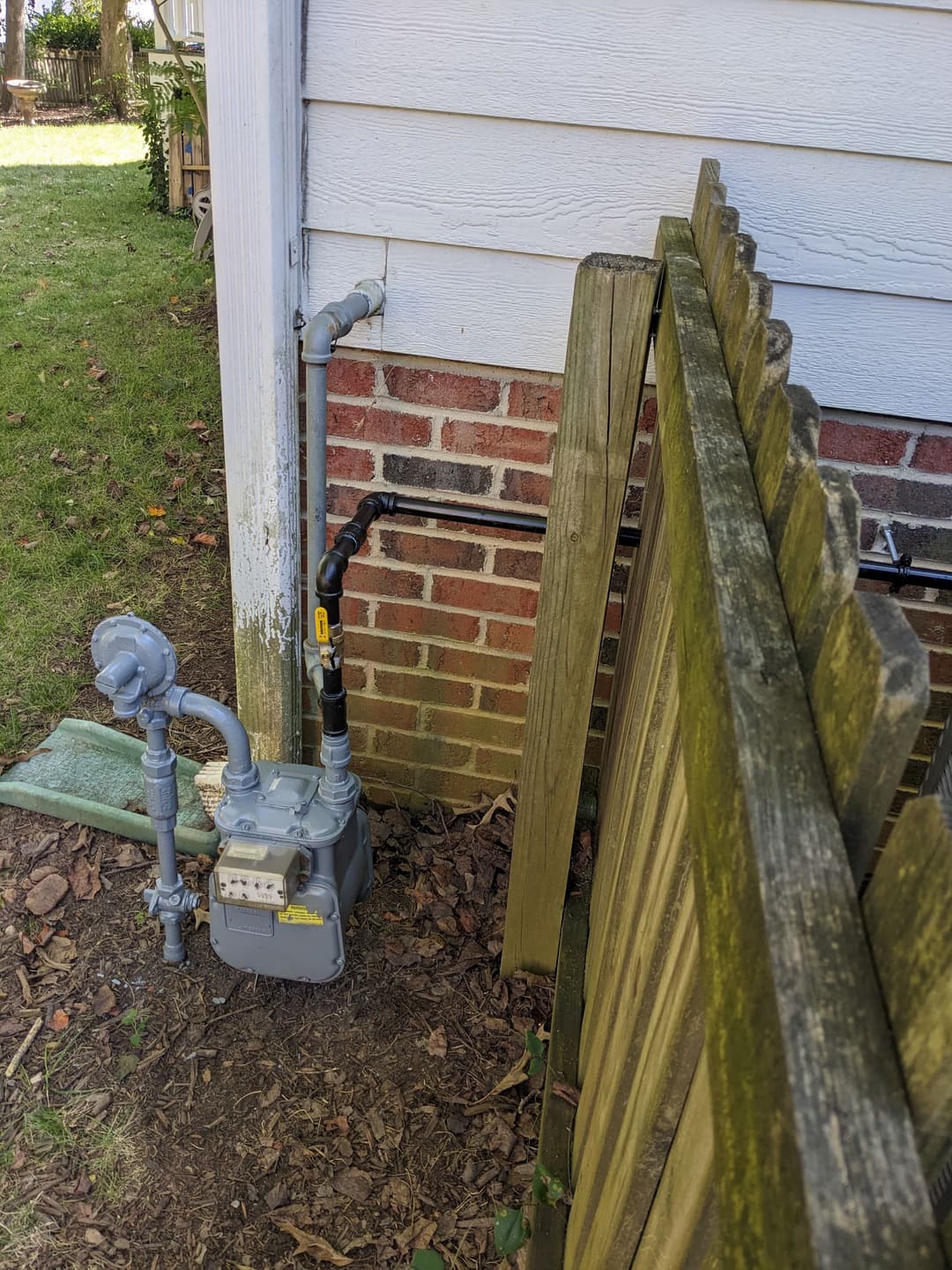 Gas meter installed on the side of a house next to a wooden fence and plumbing connections.